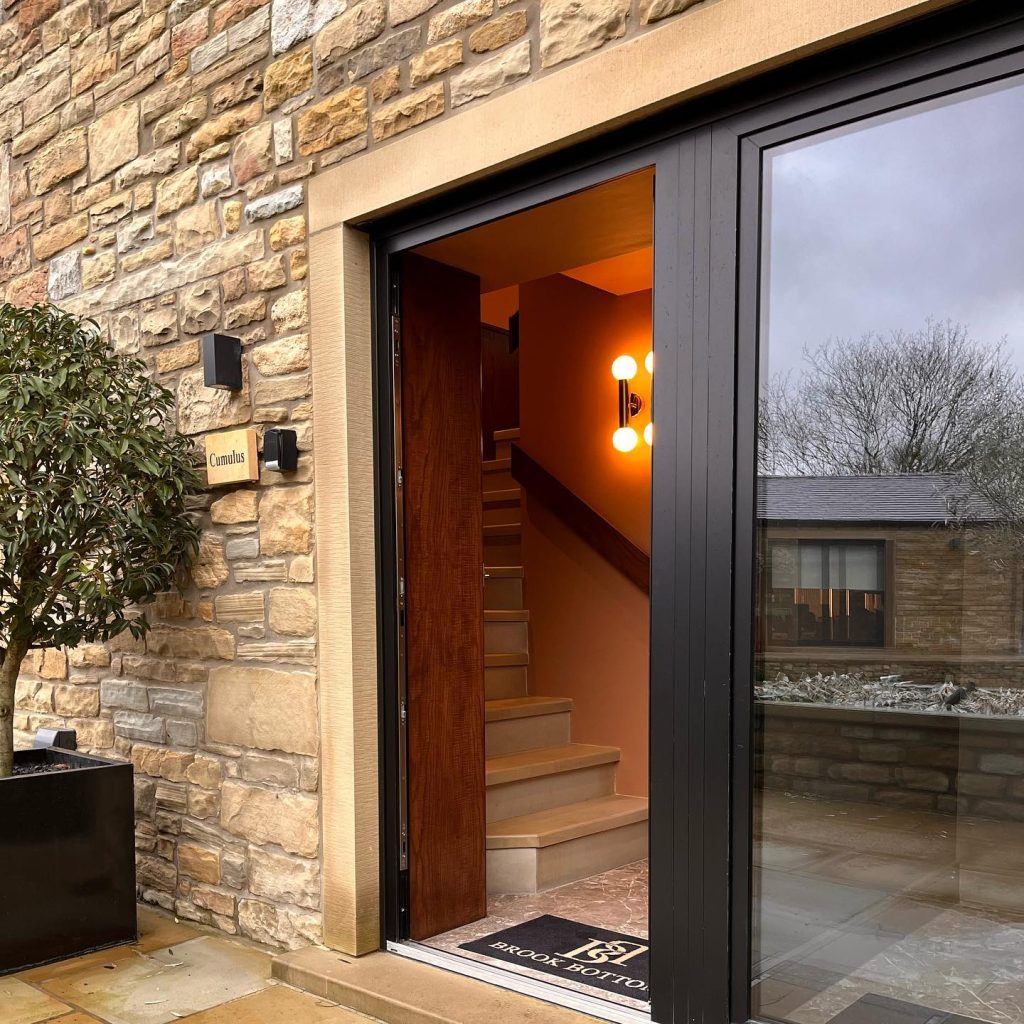 Open doorway of a modern home showing warm interior lighting and a wooden staircase, framed by black aluminium door frames, set in a natural stone wall with a 'Cumulus' nameplate
