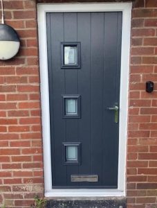 A black composite front door with three square glass panels, fitted within a white door frame against a red brick wall, under an outdoor lamp.
