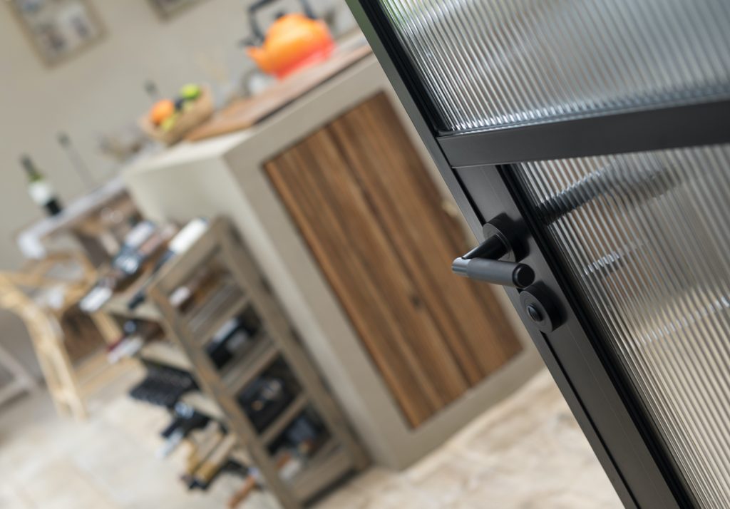 Close-up of a black aluminium door handle and frosted glass panel, with a softly focused background featuring a kitchen interior with wooden furnishings.