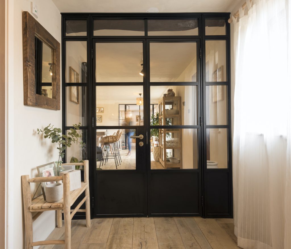 Chic interior view through black aluminium framed glass doors, showing a warmly lit, cosy room with wooden furniture and a welcoming, modern décor.