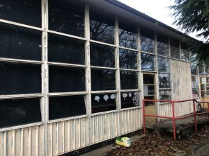 Exterior view of an old school building showing worn and peeling white window frames set in a concrete wall, with a red handrail leading to a doorway, indicating a need for renovation.