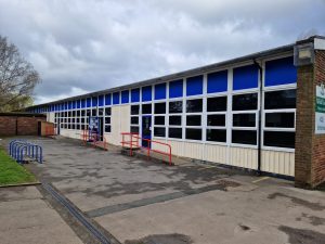 Refurbished school building with new blue and white UPVC windows and doors, and a red handrail on the entrance ramp, set against a cloudy sky.