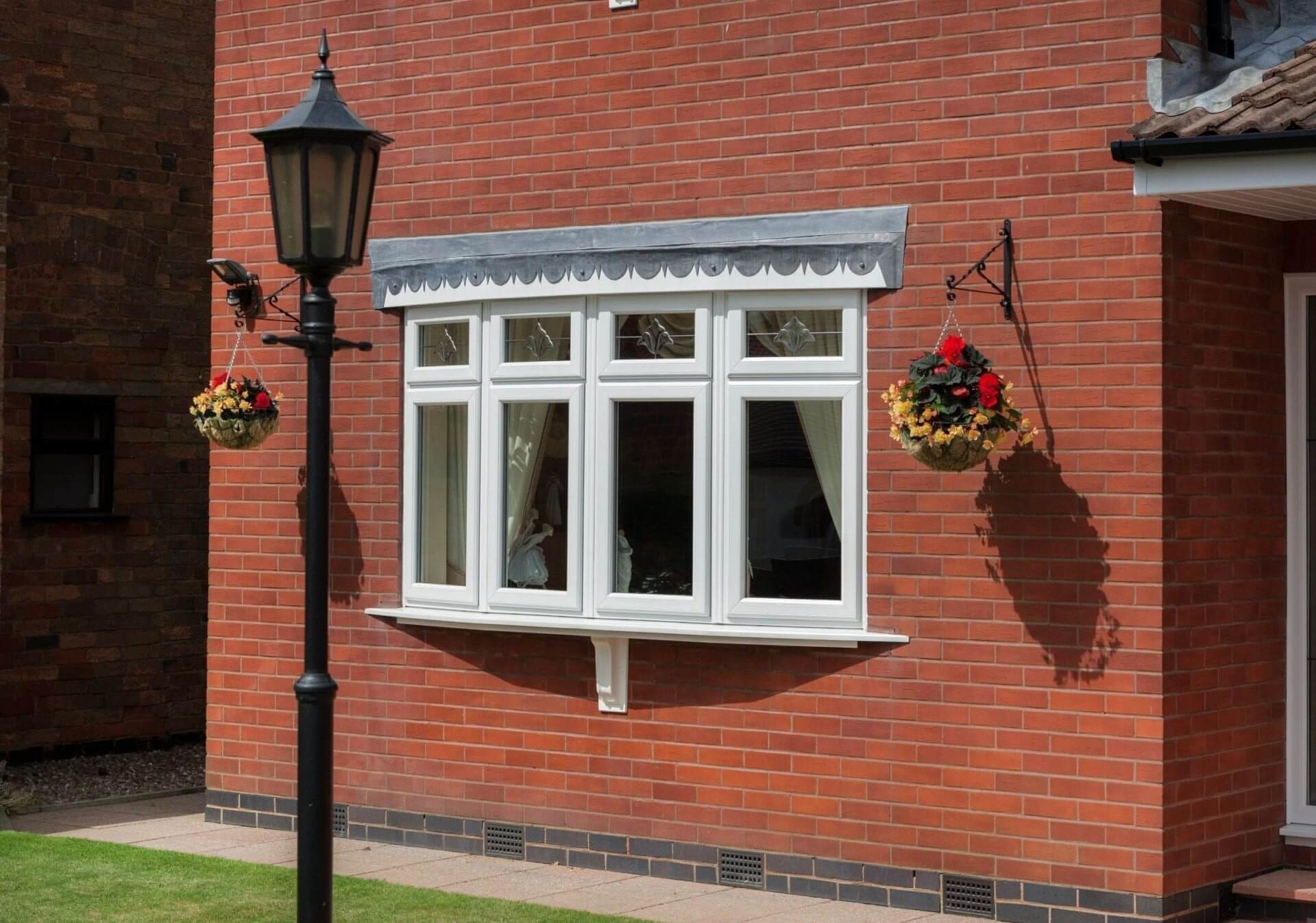 White uPVC bay and bow window with decorative glazing bars on a red brick house, complemented by hanging flower baskets and a traditional outdoor lantern.