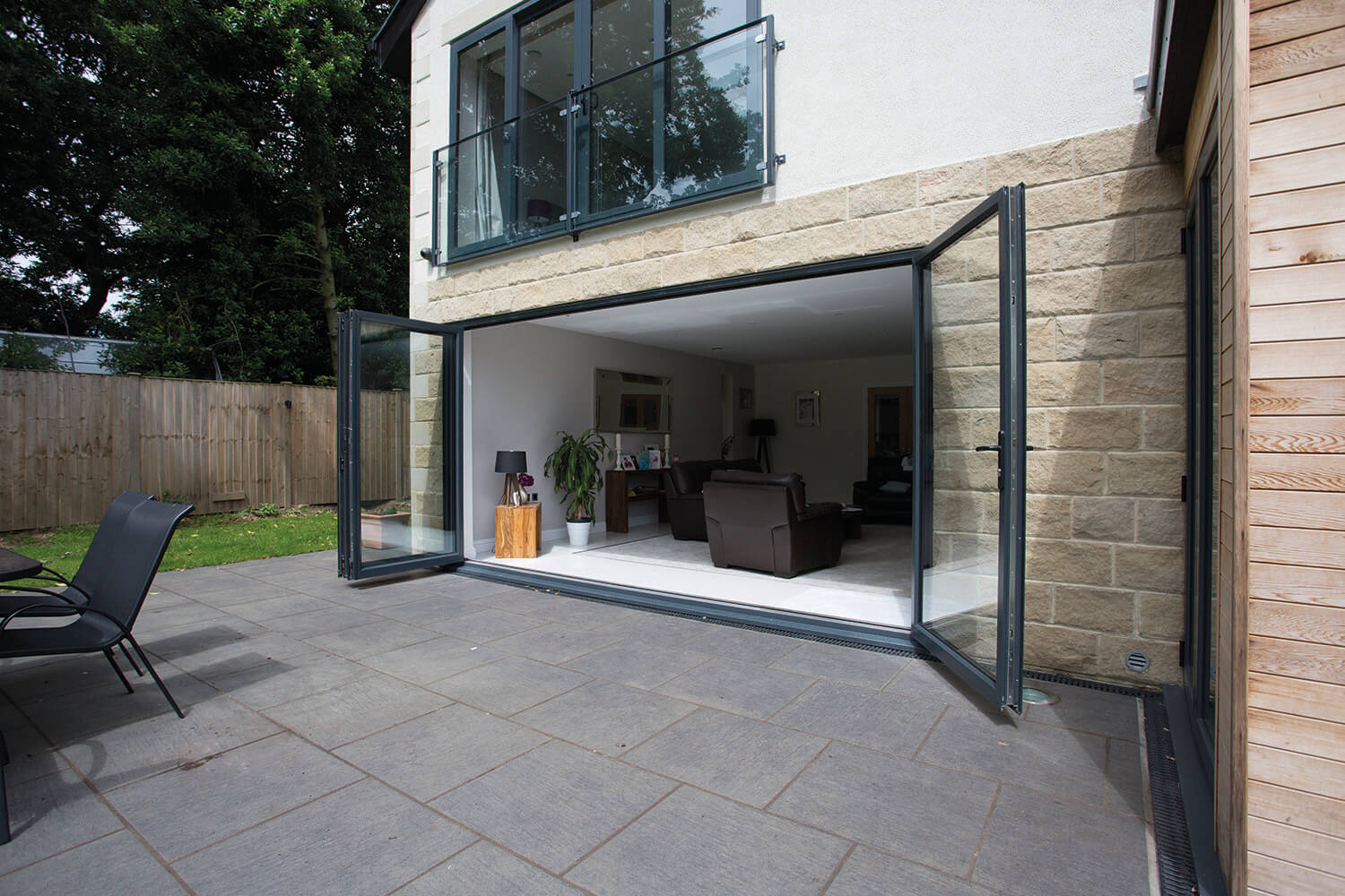 Spacious and modern living area extended outdoors with fully opened energy-efficient aluminium bifold doors, leading to a paved patio in a serene garden setting.