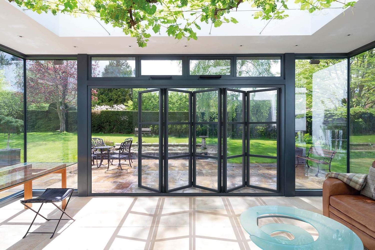 A sophisticated and modern living space featuring heritage-style aluminium bi-folding doors that open to a lush garden. The doors have a sleek black frame with multiple glass panes, offering a clear view of the green lawn and flowering trees outside. Natural light floods the room, casting soft shadows on the cream tiled floor. The interior is furnished with a brown leather sofa and a unique blue glass coffee table, with a black metal chair to the side, creating a seamless blend of comfort and contemporary design.