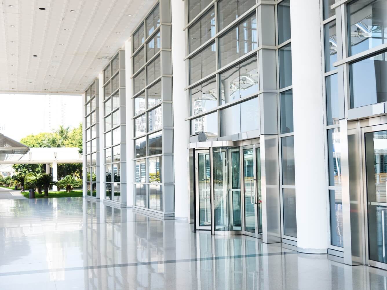 Interior view of a modern building lobby featuring floor-to-ceiling glass curtain walls with metal frames, allowing ample natural light to illuminate the glossy tiled floor. Revolving glass doors provide entrance, with potted plants and city buildings visible outside.